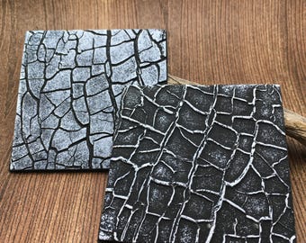 May include: Two square coasters featuring a high-contrast crackle pattern. One coaster displays a light grey surface with deep black fissures, while the other features a solid black surface with prominent white raised crackle lines.