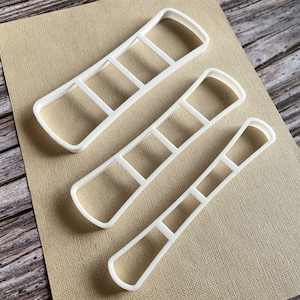 May include: Three white plastic cookie cutters shaped like ladders, each with a different size. The cutters are on a brown textured surface.