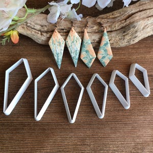 May include: Four diamond-shaped earrings featuring a marbled pattern of peach and sage green colors, accompanied by six white plastic diamond-shaped cutters of varying sizes. The earrings have bronze-toned metal hooks for attachment.