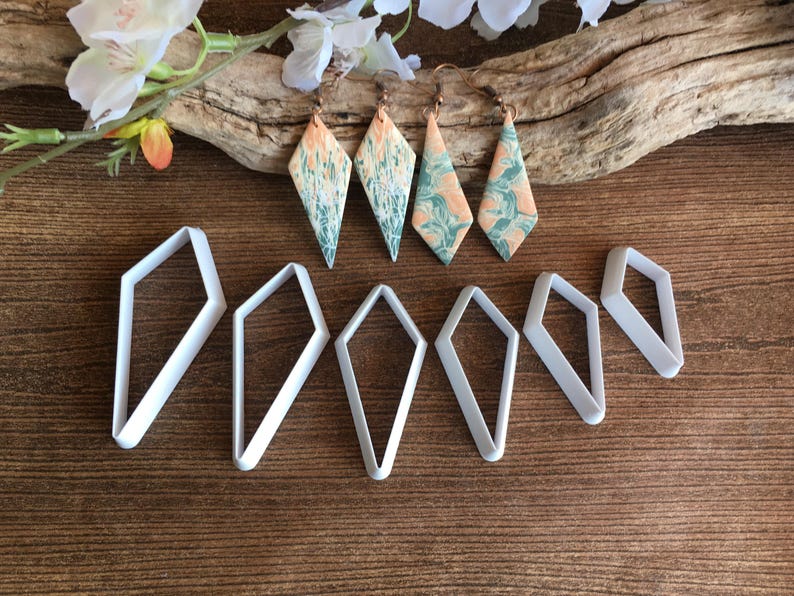 May include: Four diamond-shaped earrings featuring a marbled pattern of peach and sage green colors, accompanied by six white plastic diamond-shaped cutters of varying sizes. The earrings have bronze-toned metal hooks for attachment.