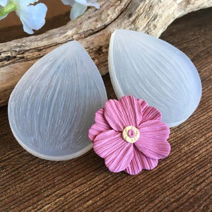 Pode incluir: Duas formas de silicone transparentes em forma de gota com detalhes texturizados, ao lado de uma flor de papel rosa com centro amarelo. Os itens estão dispostos em uma superfície de madeira, sugerindo um tema de artesanato ou faça você mesmo.