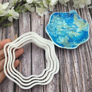 May include: Set of four white metal cookie cutters shaped like a flower with wavy edges. The cutters are nested inside each other.