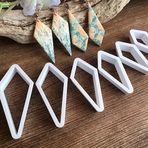 May include: Six white plastic diamond-shaped cookie cutters on a wooden surface. Three pairs of earrings with a marbled pattern in shades of green and orange are also visible.