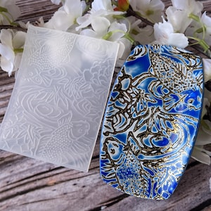 May include: A white silicone mold with a raised design of koi fish and water plants. The mold is next to a blue and gold ceramic dish with a similar design.