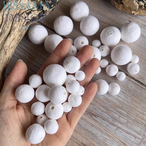 May include: A collection of white, round beads of varying sizes, some with holes, are held in a hand. The beads have a textured surface and appear to be made of a fibrous material. The background is a light-colored wooden surface.
