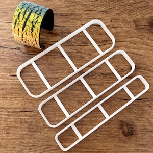 May include: Three white rectangular cookie cutters with rounded corners and interior supports are arranged on a wooden surface. A cuff bracelet with a yellow, black, and blue pattern is in the upper left corner.