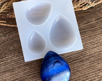May include: A square, translucent silicone mold featuring three teardrop-shaped cavities of varying sizes. Positioned below the mold is a single, polished, teardrop-shaped stone displaying vibrant shades of deep blue and black with a smooth finish.