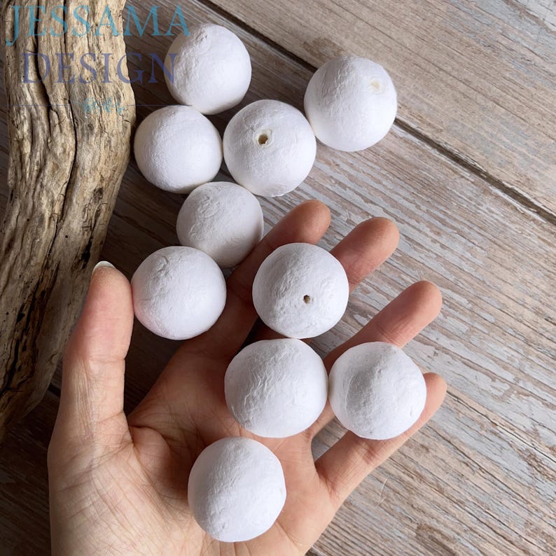 May include: A collection of white, spherical craft beads with a textured surface. Some beads have a small hole. The beads are held in a hand, with others scattered on a light brown wooden surface. The image has the text "JESSAMA DESIGN" in the upper left corner.