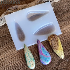 May include: A clear silicone mold with four teardrop-shaped cavities, containing three finished resin pendants in green, purple, and yellow. The pendants are next to the mold. The mold also contains two teardrop-shaped resin pieces with a wood grain appearance.