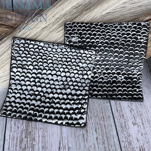 May include: Two square coasters featuring a repeating black and silver scale pattern. The design consists of rows of small, rounded, overlapping shapes that create a textured, metallic appearance across the surface of each individual coaster.