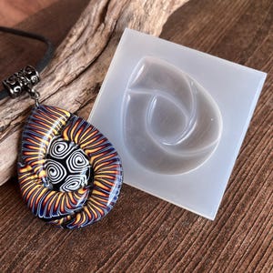 May include: A colorful glass pendant with a swirling pattern, hanging from a black cord. The pendant is shown next to a clear silicone mold with a similar swirling design.