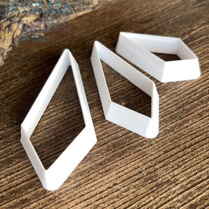 May include: Three white, diamond-shaped cutters with open centers and angled walls. The cutters are arranged in a cluster, featuring a sharp, geometric outline. The white plastic material has a smooth finish, and the shapes vary slightly in their proportions.