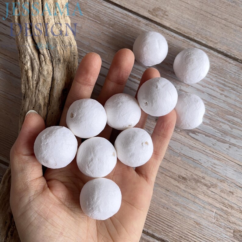 May include: A hand holds several small, white, spherical decorative balls. The balls have a textured surface and are arranged in a cluster. The background includes a piece of driftwood and a wooden surface.