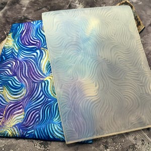 May include: A blue and purple patterned fabric next to a clear, textured rectangular mold. The fabric has a swirling design with gold accents. The mold has a similar wavy pattern. The items are displayed on a textured grey surface.
