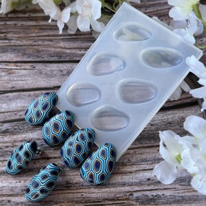 May include: A clear silicone mold with six oval-shaped cavities.  The mold is next to five blue, gold, and white patterned oval-shaped polymer clay earrings.