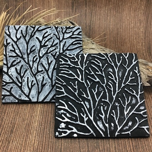 May include: Two square black and white coasters with a raised design of branches. The coasters are made of a textured material and have a rustic look.