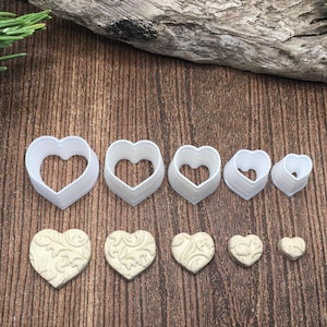 May include: A set of 7 white plastic heart-shaped cookie cutters of varying sizes, along with 5 heart-shaped cookies decorated with a swirl pattern.