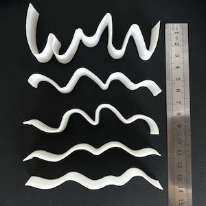 May include: Five white, wavy, 3D-printed shapes of varying lengths and curves are arranged on a black surface. A ruler with centimetre markings is positioned vertically along the right side, showing measurements up to 15 cm.