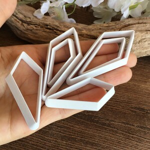May include: A set of four white plastic diamond-shaped cookie cutters of varying sizes. The cutters feature a hollow center and a thin, flat rim, arranged in a cluster to display their geometric kite-like outlines and distinct angular forms.