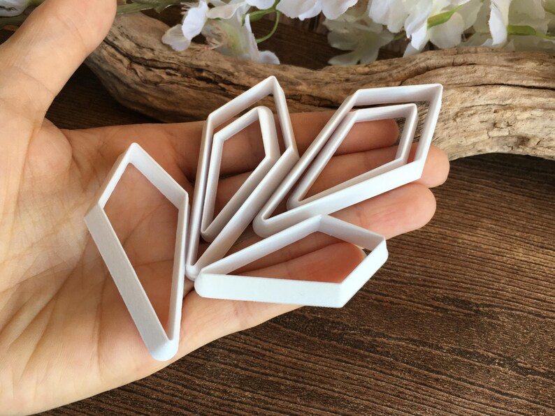 May include: A set of four white plastic diamond-shaped cookie cutters of varying sizes. The cutters feature a hollow center and a thin, flat rim, arranged in a cluster to display their geometric kite-like outlines and distinct angular forms.