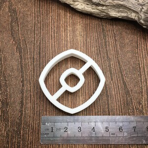 以下が含まれることがあります： 茶色の木製の表面に置かれた、白いプラスチック製のクッキーカッター。ポケボールの形で、中央に赤と白の円があります。