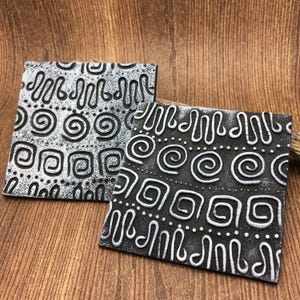 May include: Two square ceramic coasters with a black and white geometric pattern. The coasters have a raised, textured design.