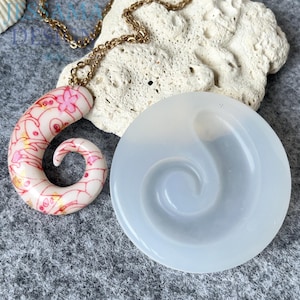 Può includere: Una collana con una catena dorata e un ciondolo a forma di spirale bianco, rosa e oro. Accanto, uno stampo in silicone circolare con un design a spirale abbinato. Il ciondolo ha un motivo floreale.