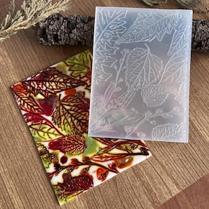 Peut inclure: Un moule en silicone transparent avec un motif de feuilles détaillé à côté d'une œuvre d'art colorée sur le thème des feuilles. L'œuvre d'art présente des feuilles rouges, jaunes et marron sur un fond blanc. Le moule est rectangulaire et montre diverses formes de feuilles.