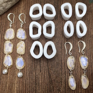 May include: Two pairs of dangle earrings with iridescent white and brown stones. Each earring has three stones and a pearl at the bottom. The earrings are displayed next to eight white rubber earring backs.