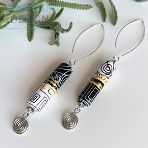May include: A pair of dangle earrings featuring cylindrical beads with black and white geometric patterns and gold-colored accents. Each earring hangs from a silver-toned curved wire and terminates in a small, flat, silver-toned spiral charm.