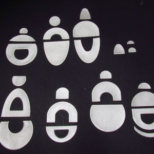 May include: A set of nine white, geometric shapes, including circles, ovals, and arches, cut from a reflective material. The shapes are arranged in a random pattern on a black background.