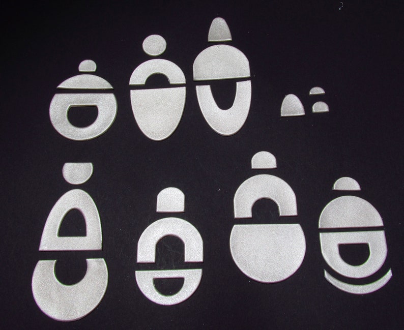 May include: A set of nine white, geometric shapes, including circles, ovals, and arches, cut from a reflective material. The shapes are arranged in a random pattern on a black background.