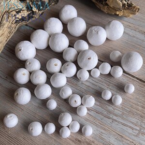 May include: A collection of white, spherical beads of varying sizes, scattered on a wooden surface. Each bead has a small hole, suggesting they are for stringing. The beads are made of a textured material, possibly paper or fabric.