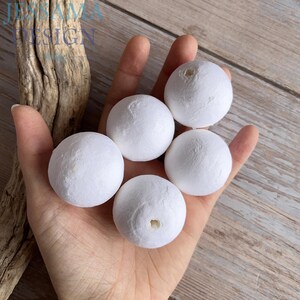 May include: Five white, spherical decorative balls with a textured surface are held in a hand. The balls are evenly spaced and appear to be made of a lightweight material. The background is a light wood surface.
