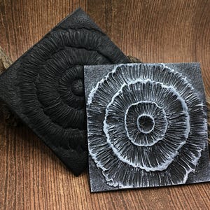 May include: Two square black and white coasters with a circular, concentric pattern. The coaster on the left is black with a raised, textured pattern. The coaster on the right is white with a raised, textured pattern.