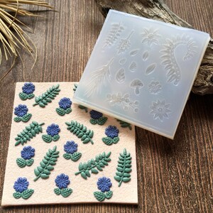 Könnte beinhalten: Eine Silikonform mit einem floralen Design, mit Blättern, Blumen und einem Pilz. Die Form wird neben einer Platte aus Polymer Clay mit blauen und grünen Blumen und Blättern gezeigt.