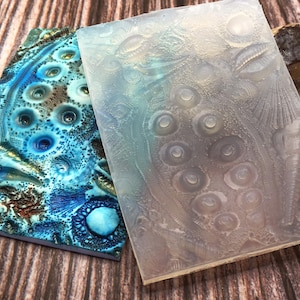 May include: Two silicone molds for crafting. One mold is blue and gold with a textured surface. The other mold is clear and has a raised design of circles and lines.