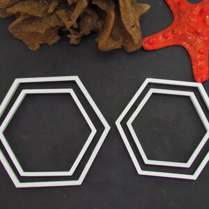 Puede incluir: Dos cortadores de galletas hexagonales de plástico blanco, uno ligeramente más grande que el otro, sobre un fondo negro.