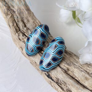 May include: Two oval-shaped earrings with a blue, brown, and white geometric pattern. The earrings are displayed on a piece of driftwood against a white background. The earrings have a glossy finish.