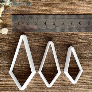 May include: Three white diamond-shaped cutters of varying sizes arranged in a row. A metal ruler is positioned above the cutters, showing the largest cutter is approximately 2.5 inches tall and the smallest is approximately 1.75 inches tall.