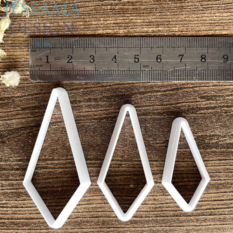 May include: Three white diamond-shaped cutters of varying sizes arranged in a row. A metal ruler is positioned above the cutters, showing the largest cutter is approximately 2.5 inches tall and the smallest is approximately 1.75 inches tall.