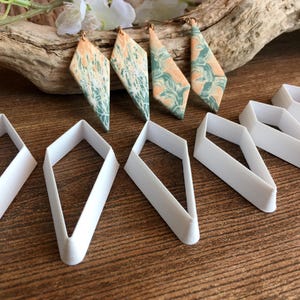 May include: Four diamond-shaped earrings featuring a marbled pattern of peach and teal colors, accompanied by five white plastic diamond-shaped cutters. The earrings have metal hooks and are displayed alongside the matching geometric tools.