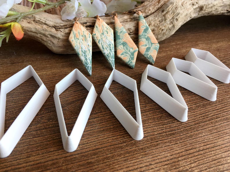 May include: Four diamond-shaped earrings featuring a marbled pattern of peach and teal colors, accompanied by five white plastic diamond-shaped cutters. The earrings have metal hooks and are displayed alongside the matching geometric tools.