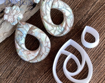 May include: A pair of iridescent teardrop earrings with a floral accent, alongside white teardrop-shaped polymer clay cutters. The earrings have a pearlescent finish with hints of blue, green, and brown. The background is a dark wood surface.