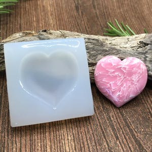 Könnte beinhalten: Eine transparente Silikonform in Herzform, neben einem rosafarbenen herzförmigen Harzstück mit Marmorierung.
