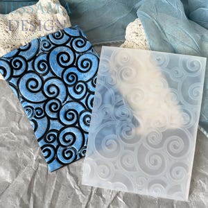 May include: Two textured embossing folders. One is blue with black swirls, the other is clear with a swirl pattern. The folders are rectangular and likely used for crafting or scrapbooking. The image has a soft, diffused lighting.
