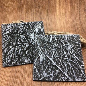 May include: Two square coasters with a textured, abstract design. The coasters are mainly black with white, vein-like patterns. The coasters are displayed on a wooden surface.