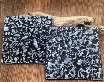 May include: Two square coasters featuring a textured, cratered surface with a high-contrast black and metallic silver finish. The design mimics a rugged, rocky terrain with deep indentations and raised, shimmering silver highlights across the top.