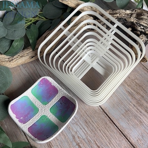 May include: A set of nested white square cutters with rounded corners and a white square tray featuring four recessed sections with a textured, iridescent purple and green finish that shifts in colour across the surface of each square section.