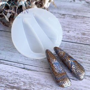 May include: A clear, circular silicone mold with two teardrop-shaped cavities, next to two decorative resin pieces. The resin pieces are elongated, with a mosaic-like pattern in shades of brown, silver, and blue. The mold is on a light wood surface.